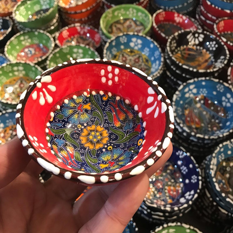 May include: A small, hand-painted ceramic bowl with a vibrant floral design. The interior features a detailed pattern of blue, yellow, and green flowers on a dark blue background. The rim is red with white accents. Other bowls in various colors are in the background.
