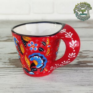 May include: A red ceramic coffee cup with a black rim and a white handle. The cup is decorated with a floral pattern in blue, yellow, and black.