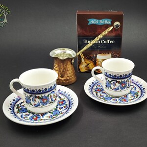 May include: Two white ceramic coffee cups and saucers decorated with blue and red floral patterns, accompanied by a metallic copper-colored coffee pot and a brown box labeled Ağa Baba Turkish Coffee with an image of a coffee cup.