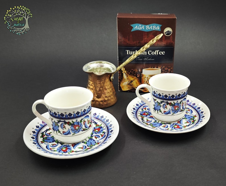 May include: Two white ceramic coffee cups and saucers decorated with blue and red floral patterns, accompanied by a metallic copper-colored coffee pot and a brown box labeled Ağa Baba Turkish Coffee with an image of a coffee cup.