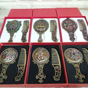 以下が含まれることがあります： 赤いギフトボックスに入った、アンティークスタイルの手鏡と櫛。鏡と櫛には、カラフルな宝石のアクセントが付いた、複雑な花柄と孔雀のデザインが施されています。金属はブロンズ仕上げです。セットは一列に並んでいます。