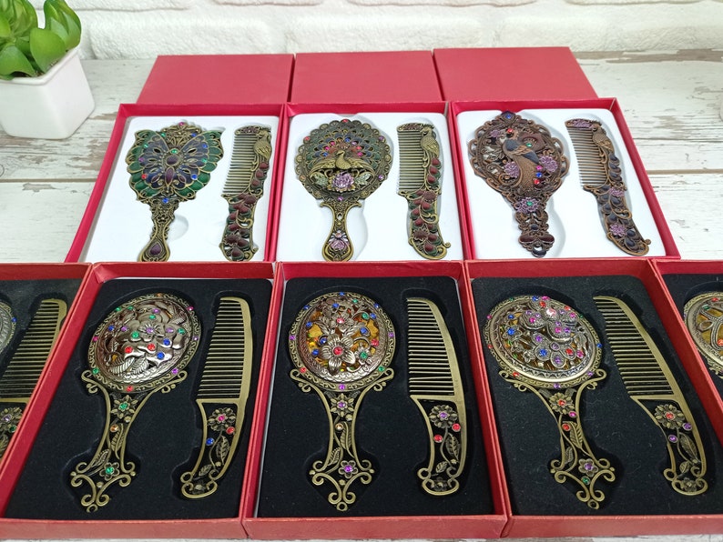 May include: Ornate antique-style hand mirrors and combs in red gift boxes. The mirrors and combs have intricate floral and peacock designs with colorful jewel accents. The metal has a bronze finish. The sets are displayed in a row.