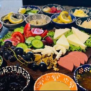 May include: A wooden serving board laden with a variety of breakfast foods. Includes sliced tomatoes, cucumbers, cheese, grapes, walnuts, and sliced sausage. Small bowls of jam, olives, and other condiments surround the board. The bowls have colorful patterns.