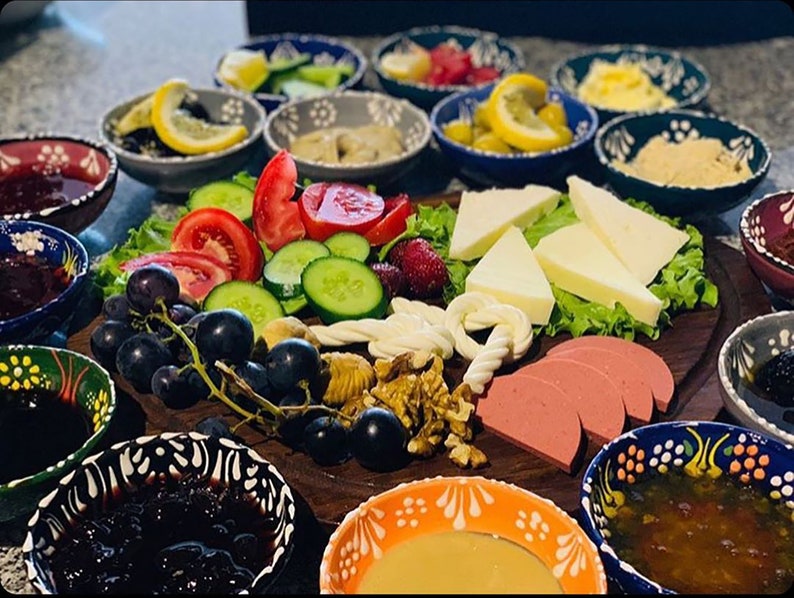 May include: A wooden serving board laden with a variety of breakfast foods. Includes sliced tomatoes, cucumbers, cheese, grapes, walnuts, and sliced sausage. Small bowls of jam, olives, and other condiments surround the board. The bowls have colorful patterns.