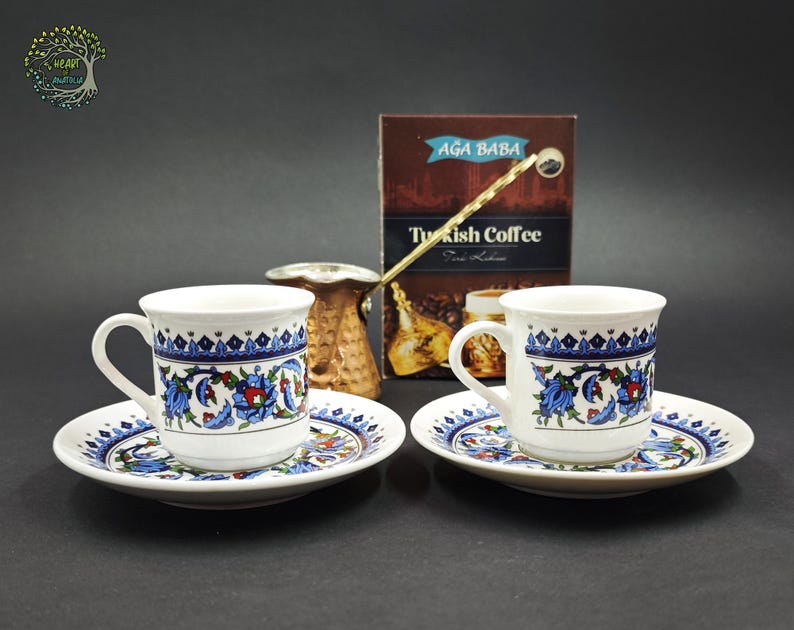May include: Two white ceramic coffee cups and matching saucers featuring blue, red, and green floral patterns. A copper-colored metal coffee pot sits behind the cups, next to a brown box labeled AGA BABA Turkish Coffee with a graphic of coffee beans.