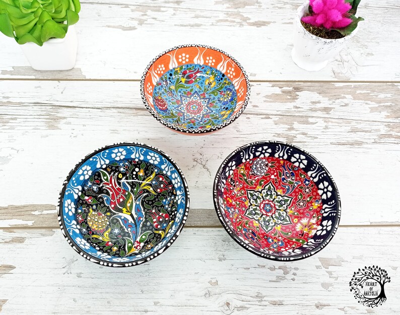 Puede incluir: Tres cuencos decorativos de cer&aacute;mica con intrincados dise&ntilde;os florales. Los cuencos presentan colores vibrantes como el rojo, el azul, el naranja y el verde. Cada cuenco tiene un dise&ntilde;o &uacute;nico, con patrones detallados sobre un fondo negro o blanco. Los cuencos est&aacute;n sobre una superficie de madera blanca.