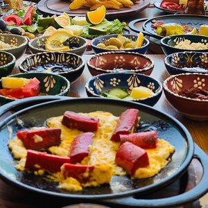 May include: A table laden with a variety of breakfast foods. A black skillet holds scrambled eggs topped with red sausage slices. Small, decorative bowls contain olives, jams, and other side dishes. Sliced tomatoes, oranges, and other fruits and vegetables are also present.