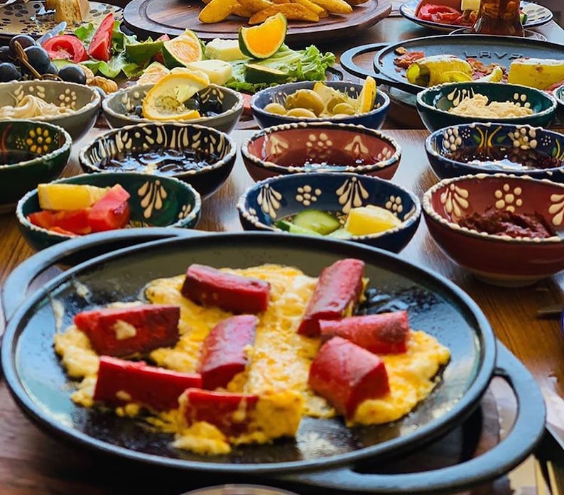 May include: A table laden with a variety of breakfast foods. A black skillet holds scrambled eggs topped with red sausage slices. Small, decorative bowls contain olives, jams, and other side dishes. Sliced tomatoes, oranges, and other fruits and vegetables are also present.