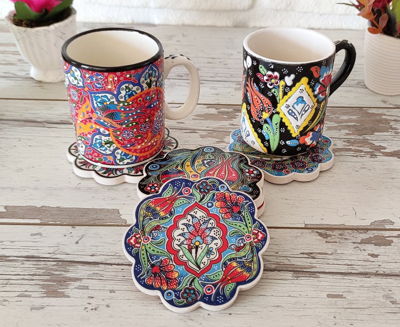 Puede incluir: Dos tazas de cer&aacute;mica con intrincados y coloridos dise&ntilde;os florales descansan sobre posavasos a juego. Los posavasos y las tazas presentan una vibrante mezcla de dise&ntilde;os rojos, azules, amarillos y verdes. Las tazas tienen bordes y asas negros, y los posavasos tienen un borde festoneado.
