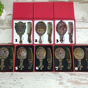May include: Ornate antique-style vanity mirror and comb sets in red gift boxes. The mirrors and combs have intricate designs with colorful jewel accents. The sets are displayed on a light wood surface.