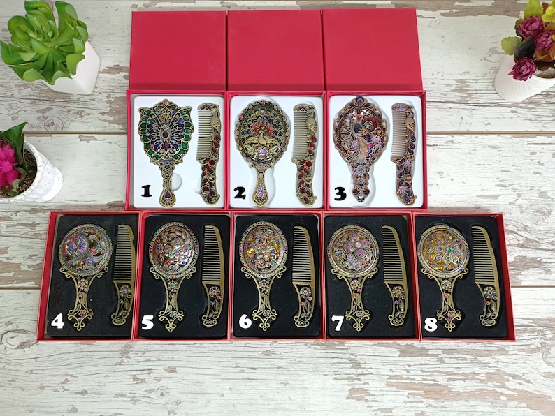 May include: Ornate antique-style vanity mirror and comb sets in red gift boxes. The mirrors and combs have intricate designs with colorful jewel accents. The sets are displayed on a light wood surface.