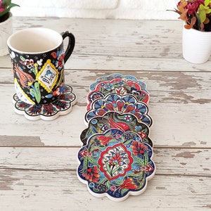 Puede incluir: Un juego de posavasos de cer&aacute;mica con motivos florales coloridos y borde festoneado. Los posavasos presentan un dise&ntilde;o vibrante con elementos rojos, azules y verdes. Una taza a juego descansa sobre uno de los posavasos. El fondo es una superficie de madera blanca.