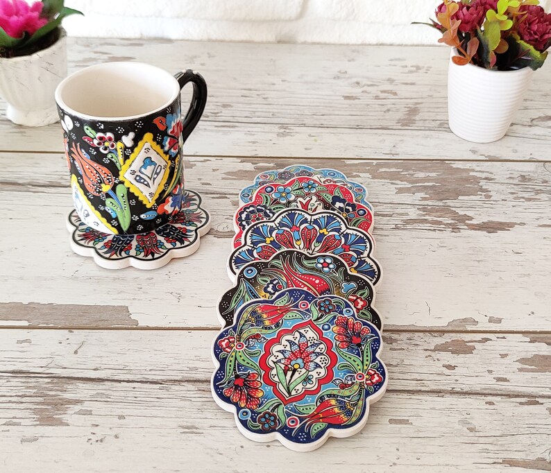 Puede incluir: Un juego de posavasos de cer&aacute;mica con motivos florales coloridos y borde festoneado. Los posavasos presentan un dise&ntilde;o vibrante con elementos rojos, azules y verdes. Una taza a juego descansa sobre uno de los posavasos. El fondo es una superficie de madera blanca.