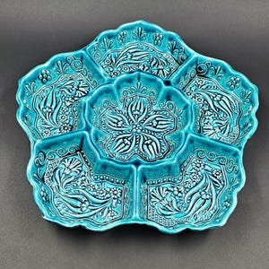 May include: A turquoise ceramic serving dish with a floral design. The dish is divided into six sections, each with a detailed pattern of flowers and leaves. The edges of the dish are scalloped.