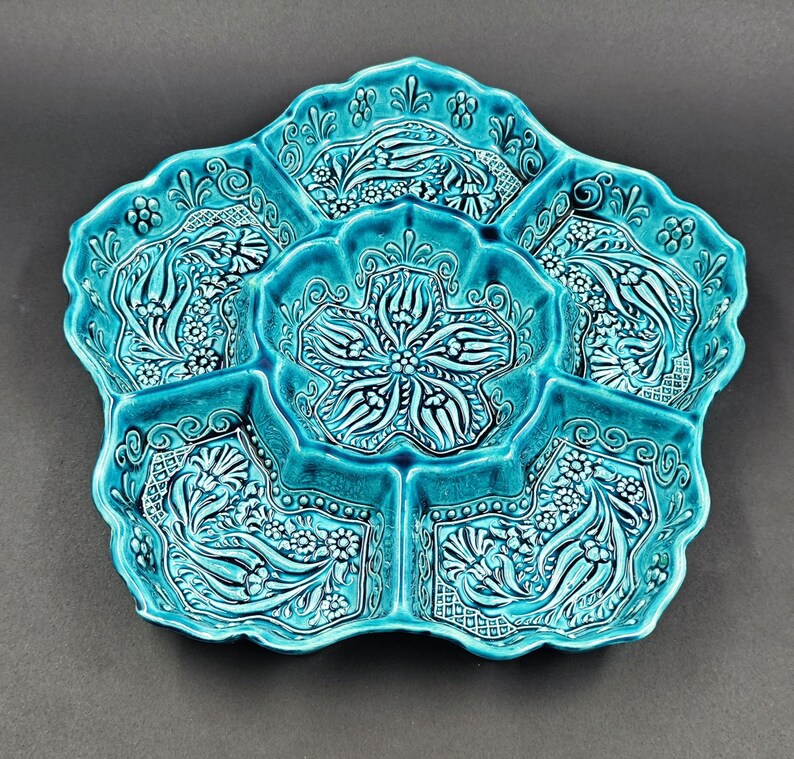 May include: A turquoise ceramic serving dish with a floral design. The dish is divided into six sections, each with a detailed pattern of flowers and leaves. The edges of the dish are scalloped.