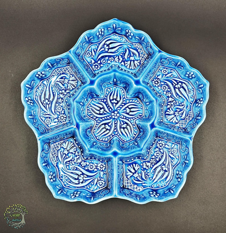 May include: A turquoise ceramic serving dish with a floral design. The dish is shaped like a flower with five sections and a central compartment. The raised white floral pattern adds a decorative touch to the serving dish.