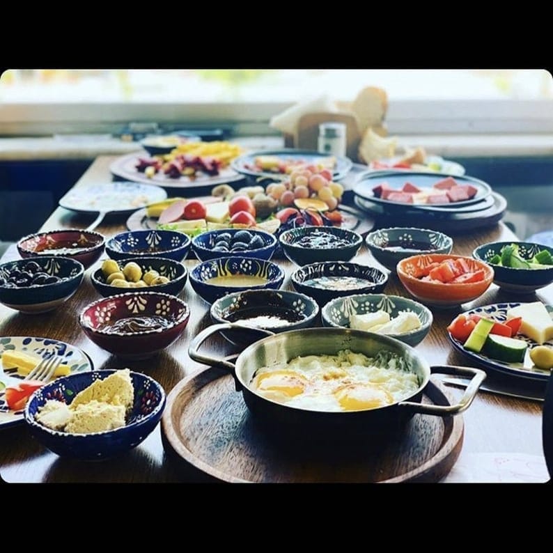 May include: A table laden with a variety of breakfast foods. Small bowls of olives, jams, and spreads surround a pan of fried eggs. Sliced meats, cheeses, and fresh fruit are also present, creating a colorful and appetizing spread.