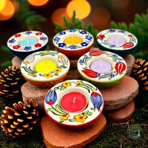 Puede incluir: Seis portavelas de cerámica coloridos con diseños florales y una vela de té en cada uno. Los portavelas están dispuestos en una pila de rocas con piñas a cada lado.