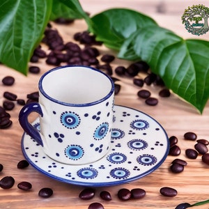 May include: A white ceramic coffee cup and saucer with a blue and white eye pattern. The cup and saucer are on a wooden surface with coffee beans scattered around them.