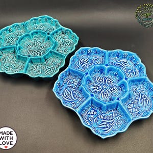 May include: Two ceramic serving platters in shades of blue and turquoise. Each platter has a central compartment surrounded by six smaller sections. The platters feature a floral and leaf design in white relief. The platters are handmade and labeled "MADE WITH LOVE".