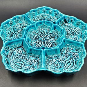 May include: A turquoise ceramic serving dish with a floral design. The dish is divided into five sections, each with a detailed, raised pattern. The edges are scalloped, and the overall shape is reminiscent of a flower. The dish is likely handmade.