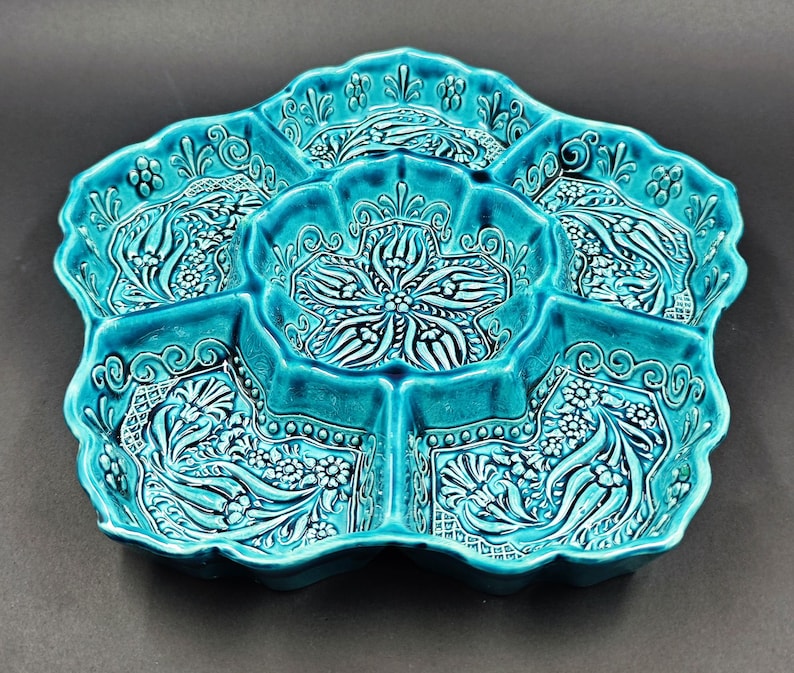 May include: A turquoise ceramic serving dish with a floral design. The dish is divided into five sections, each with a detailed, raised pattern. The edges are scalloped, and the overall shape is reminiscent of a flower. The dish is likely handmade.