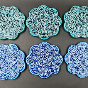 May include: Six ceramic coasters with a floral design. The coasters are in shades of blue and turquoise, with intricate white patterns. Each coaster has a scalloped edge and measures approximately 4 inches in diameter.