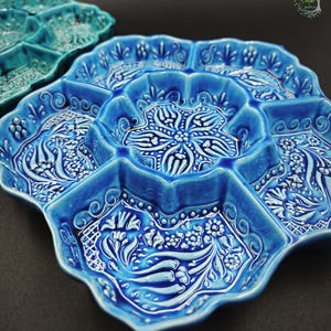 May include: A close-up of a blue ceramic serving dish with multiple compartments. The dish has a scalloped edge and is decorated with white floral and geometric patterns. Another teal dish is partially visible in the background.