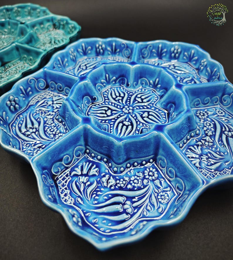 May include: A close-up of a blue ceramic serving dish with multiple compartments. The dish has a scalloped edge and is decorated with white floral and geometric patterns. Another teal dish is partially visible in the background.