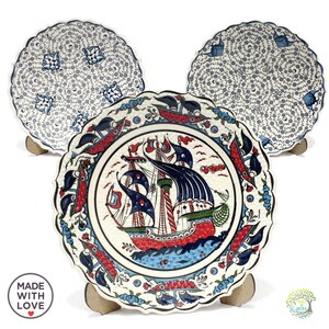 May include: Three decorative ceramic plates with a white background and blue, red, and green floral patterns. The center plate features a detailed illustration of a ship with sails.