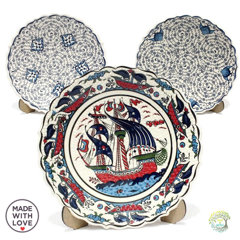 May include: Three decorative ceramic plates with a white background and blue, red, and green floral patterns. The center plate features a detailed illustration of a ship with sails.