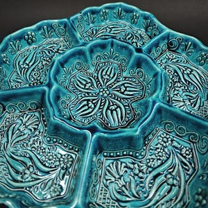 May include: A turquoise ceramic serving dish with multiple compartments. The dish features an intricate floral and geometric pattern in a raised relief design. The edges are scalloped, and the overall aesthetic is ornate and decorative.