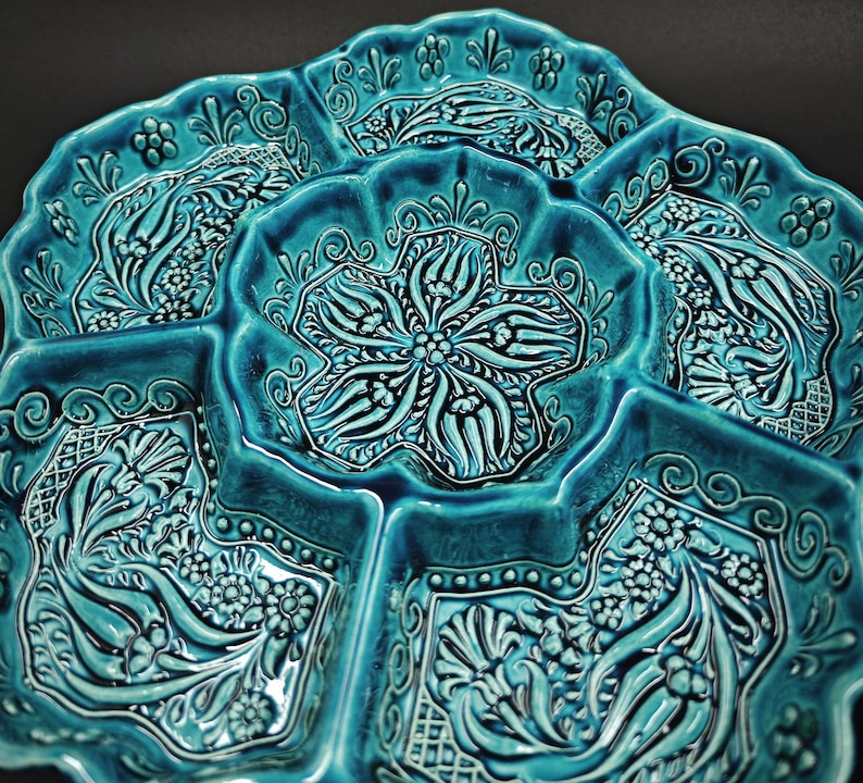 May include: A turquoise ceramic serving dish with multiple compartments. The dish features an intricate floral and geometric pattern in a raised relief design. The edges are scalloped, and the overall aesthetic is ornate and decorative.
