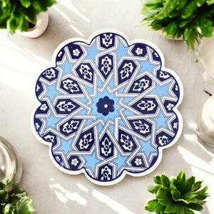 Puede incluir: Un posavasos decorativo azul y blanco con un diseño floral en forma de estrella. El posavasos tiene un borde festoneado y un motivo floral central. El intrincado patrón presenta formas geométricas sobre un fondo azul claro.