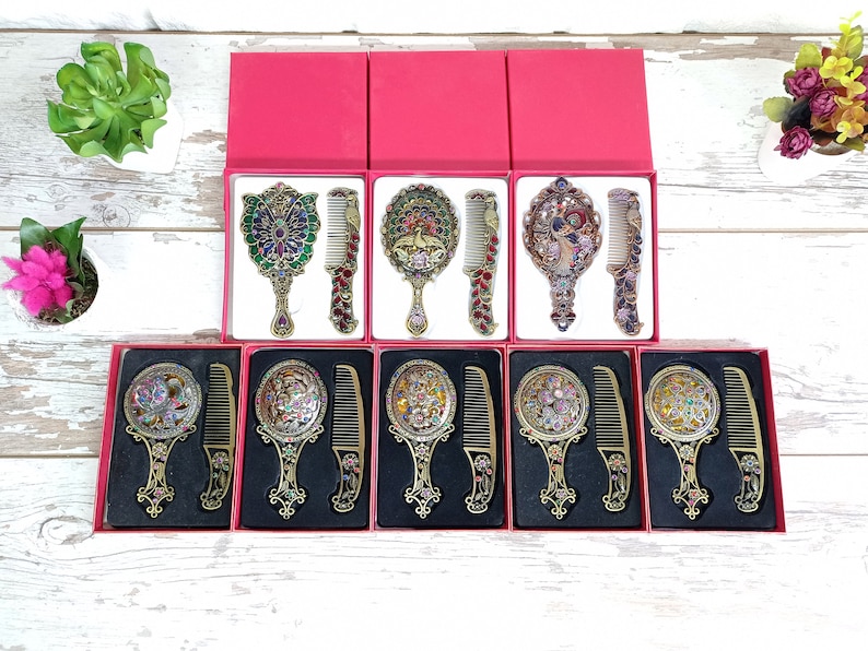 May include: Ornate vanity mirror and comb sets in red boxes. The mirrors and combs have intricate designs with colorful jewel accents. The sets are displayed on a white wooden surface, suggesting a vintage or decorative theme.