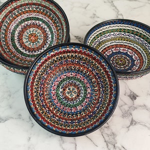 May include: A set of four decorative ceramic bowls with a black rim. Each bowl features a vibrant, intricate pattern in shades of red, blue, green, and orange, creating a mandala-like design. The bowls are arranged on a marble surface.