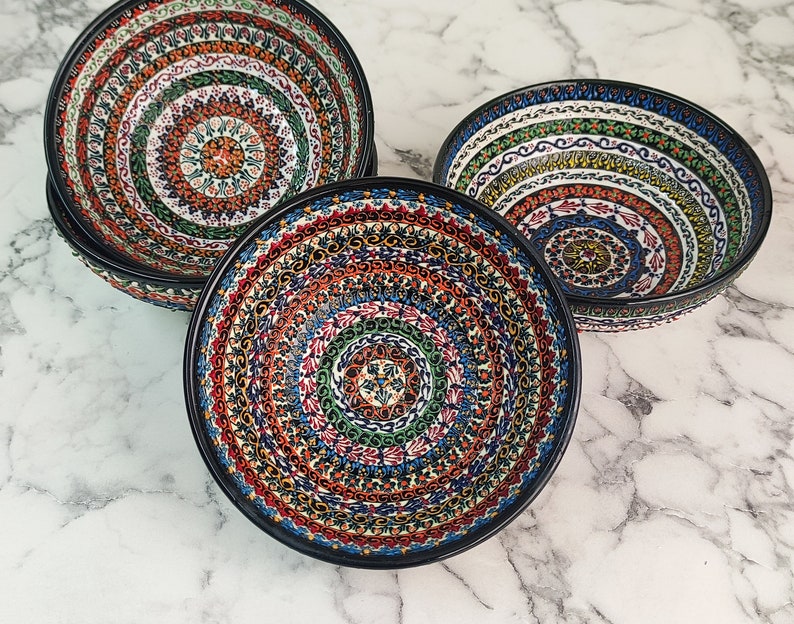 May include: A set of four decorative ceramic bowls with a black rim. Each bowl features a vibrant, intricate pattern in shades of red, blue, green, and orange, creating a mandala-like design. The bowls are arranged on a marble surface.