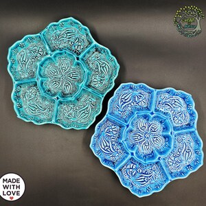 May include: Two ceramic serving platters with floral designs. One is turquoise, the other is blue. Each platter has a central flower-shaped compartment surrounded by six petal-shaped sections. The platters are decorated with white floral patterns.