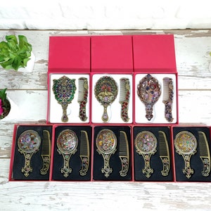 May include: Ornate vanity mirror and comb sets in red boxes. The sets feature decorative handles and frames with colorful jewel-like accents. The combs and mirrors have matching designs, creating a cohesive and elegant look. The sets are displayed on a white wooden surface.