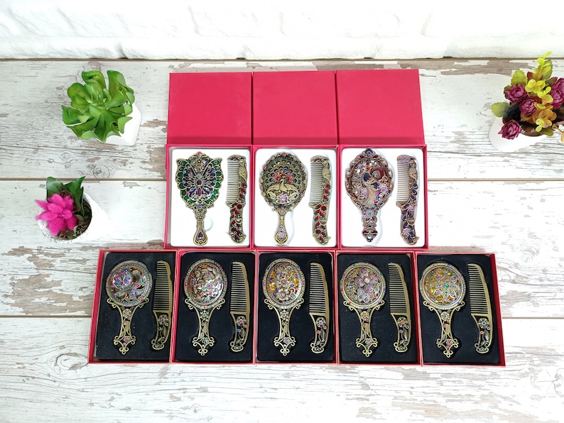 May include: Ornate vanity mirror and comb sets in red boxes. The sets feature decorative handles and frames with colorful jewel-like accents. The combs and mirrors have matching designs, creating a cohesive and elegant look. The sets are displayed on a white wooden surface.