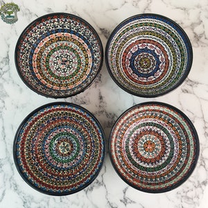 May include: Four decorative ceramic bowls with intricate, colorful patterns. Each bowl features a black rim and a unique design with concentric circles of red, blue, green, and orange. The bowls are arranged on a white marble surface.