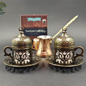 May include: Two metallic coffee cups with ornate lids and matching saucers, featuring a dark bronze finish and white inserts. A copper-colored coffee pot with a long handle sits between them, positioned in front of a box labeled AĞA BABA Turkish Coffee.