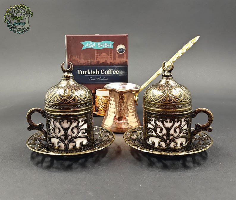 May include: Two metallic coffee cups with ornate lids and matching saucers, featuring a dark bronze finish and white inserts. A copper-colored coffee pot with a long handle sits between them, positioned in front of a box labeled AĞA BABA Turkish Coffee.
