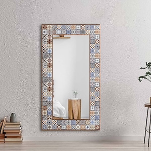May include: A full-length mirror with a decorative frame made of colorful patterned tiles. The frame is a mix of brown, blue, and white tiles. The mirror is hanging on a light gray wall.