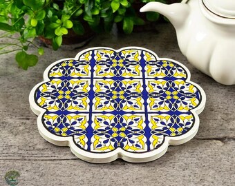 May include: A decorative, white-edged coaster with a floral tile design. The coaster features a pattern of yellow and blue floral motifs on a white background. The coaster is shaped like a stylized flower.
