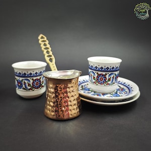 May include: A copper-colored hammered metal coffee pot with a long, decorative handle, accompanied by two white ceramic cups and saucers featuring a blue, red, and green floral pattern. The set is arranged on a dark, neutral surface.