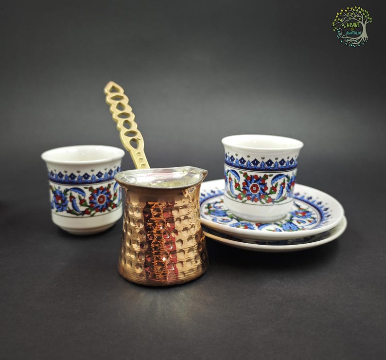 May include: A copper-colored hammered metal coffee pot with a long, decorative handle, accompanied by two white ceramic cups and saucers featuring a blue, red, and green floral pattern. The set is arranged on a dark, neutral surface.