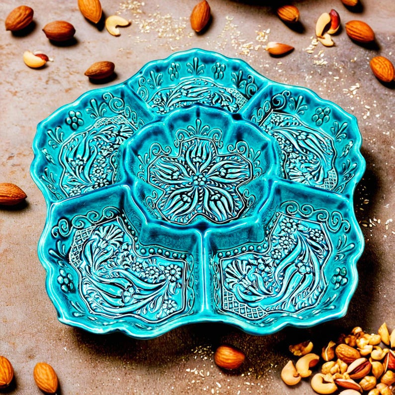 May include: A turquoise ceramic serving dish with a floral design. The dish is divided into six sections and is surrounded by almonds and cashews. The dish has a scalloped edge and is decorated with embossed floral patterns.