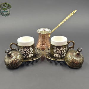 May include: A copper coffee pot with a long handle, two white ceramic cups in ornate dark metal holders, two matching saucers, and two domed lids with crescent moon finials. The set features intricate filigree patterns throughout the metalwork.