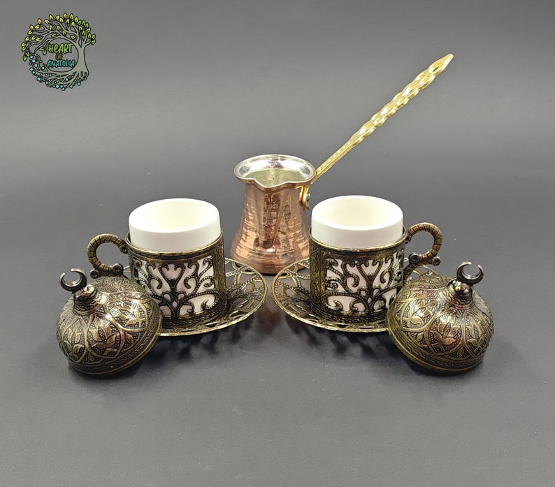May include: A copper coffee pot with a long handle, two white ceramic cups in ornate dark metal holders, two matching saucers, and two domed lids with crescent moon finials. The set features intricate filigree patterns throughout the metalwork.